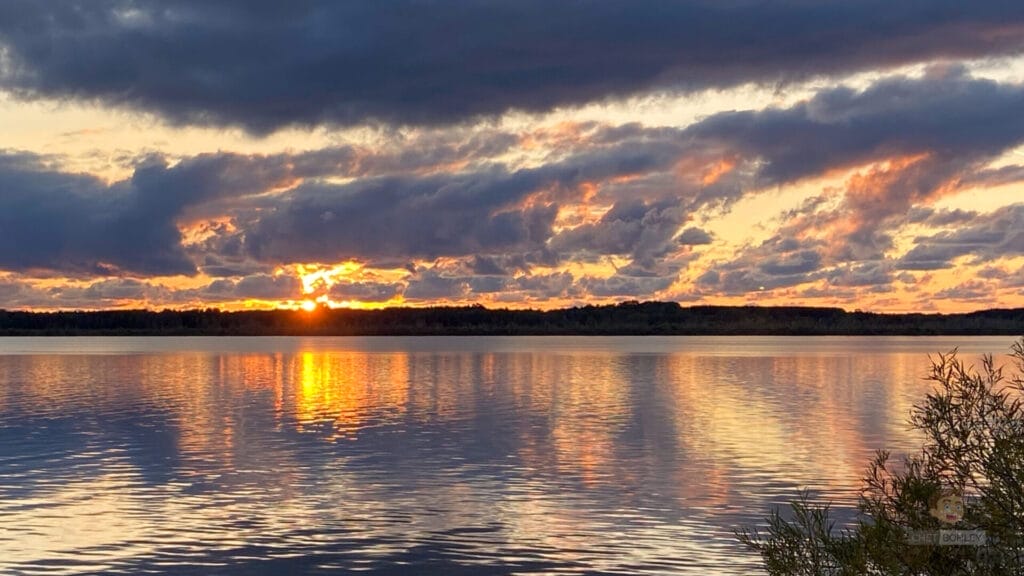 An awe-inspiring sunset over a serene lake, with fluffy clouds adding to the picturesque landscape.